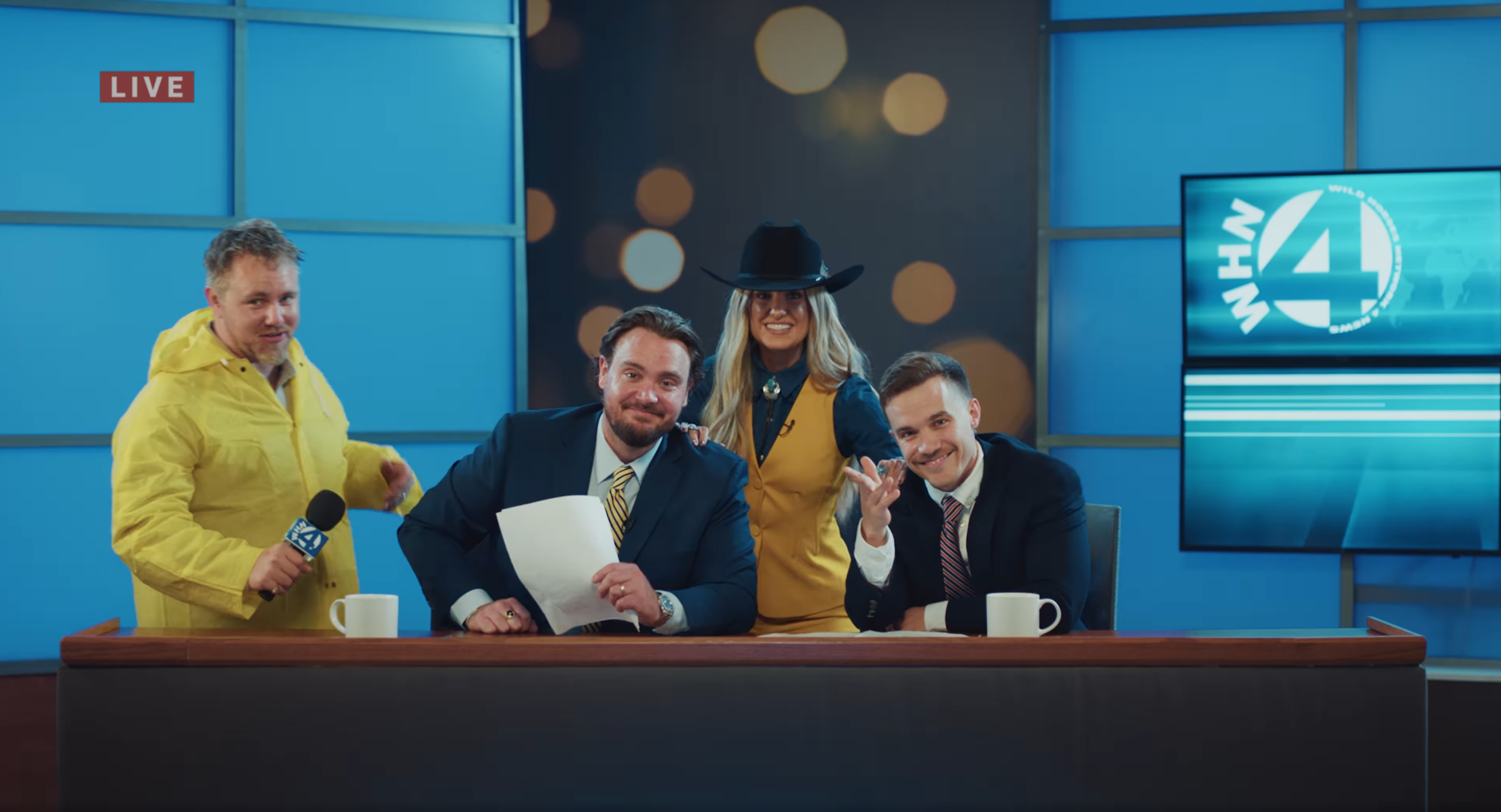 Lainey Wilson and three men dressed as news anchors pose on studio set